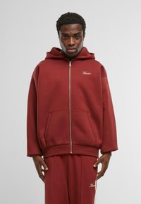 Felpa zip-up bordeaux in tessuto morbido con tasca frontale e logo ricamato. Presenta un ampio cappuccio e una vestibilità rilassata. Abbinata a pantaloni coordinati.