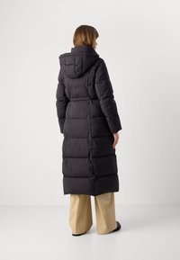 Γυναίκα που στέκεται με πλάτη, φορώντας ένα μακρύ μαύρο μπουφάν με κουκούλα, μπεζ παντελόνι σε φαρδιά γραμμή και μαύρα παπούτσια, σε απλό φόντο.