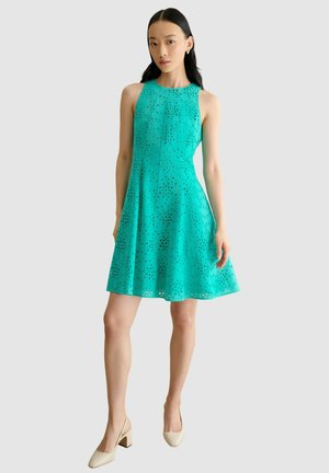 Femme aux longs cheveux noirs portant une robe turquoise sans manches avec un motif ajouré et des chaussures à talons carrés beiges, debout devant un fond uni.