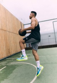 Black tank top, gray shorts, and white socks with blue and neon yellow athletic shoes. Holding a basketball, performing a jump on a court.