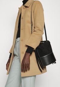 Petit sac seau noir avec texture en daim et base en cuir, doté d'une fermeture à cordon et d'une bandoulière amovible, associé à un manteau beige.