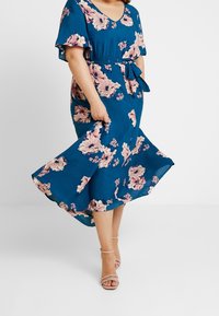 Blommig klänning i teal med korta ärmar, en midja som är insvängd och en utsvängd kjol. Lätt tyg med rosa och krämfärgade blommor samt ett knytbälte.