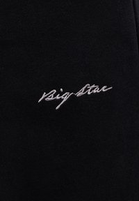 Bílá kurzíva "Big Star" vyšívaná na černé látce, zobrazující detailní texturu látky a stehu.