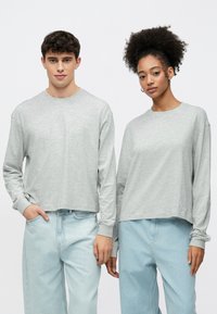 Világosszürke, hosszú ujjú, laza szabású pólók, világoskék farmerrel párosítva. Puha tapintású, kerek nyakkivágású és lejtő vállú.