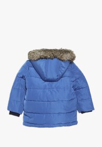 Sininen pehmustettu talvitakki hupulla, jossa on ruskea tekoturkisreuna ja mustat ribbikäänteet, nähtynä takaapäin valkoista taustaa vasten.