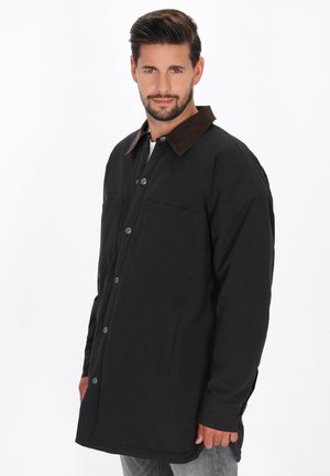 Sort jakke med en brun fløjlskrave, front snaplukninger og to store forlommer. Designet er afslappet pasform og knælangt.