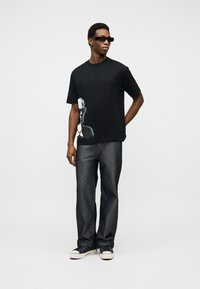 Άνδρας που φοράει μαύρο T-shirt με σχέδιο, σκούρα τζιν, μαύρα γυαλιά ηλίου και μαύρα αθλητικά παπούτσια με λευκές σόλες, στέκεται μπροστά σε ένα απλό λευκό φόντο.