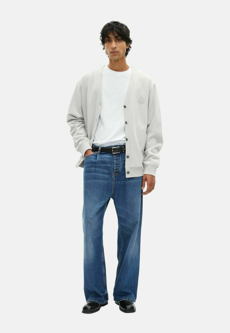 Junger Mann in einem hellgrauen, geknöpften Cardigan, weißem T-Shirt, weitem blauen Jeans mit schwarzem Gürtel und schwarzen Schuhen, der vor einem weißen Hintergrund steht.