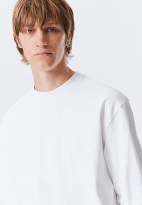 Young person with short light brown hair, wearing a white crewneck shirt and multiple earrings, against a plain light background.