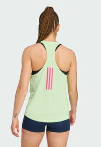 Svijetlozelena atletska majica bez rukava s dizajnom racerback, koja sadrži tri vertikalne ružičaste pruge na leđima, u kombinaciji s tamnoplavim šortsevima.