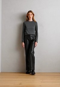 Dlhý rukáv šedý top v kombinácii s čiernymi koženými nohavicami, obe priliehavé. Model stojí na svetlej drevenej podlahe pred neutrálnou stenou.