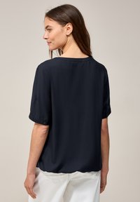 Blouse bleu marine à manches courtes avec un col rond, présentant une texture lisse et des poignets boutonnés, stylée avec un pantalon blanc.