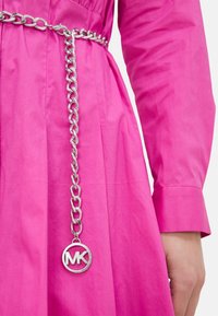Cintura in catena d'argento con accentuazione circolare del logo MK, indossata su un capo di abbigliamento di un vivace colore rosa, caratterizzato da un tessuto liscio e testurizzato.