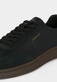 Baskets en cuir noir avec des lacets noirs, semelle marron texturée et logo "GANT" doré sur le côté.