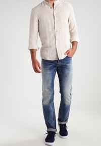 Camisa de lino color beige claro con botones y un pequeño cuello, combinada con vaqueros de denim azul deslavado y zapatillas oscuras. Estilo casual, ajuste relajado.