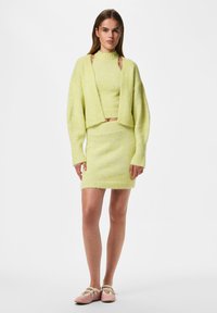 Femme portant un cardigan en maille jaune clair, un top sans manches assorti et une jupe avec des chaussures plates roses, debout devant un fond blanc uni.