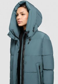 Turkusowa kurtka puchowa z dużym kapturem, zamkiem z przodu i bocznymi kieszeniami. Posiada pikowaną strukturę i regulowane troczki.