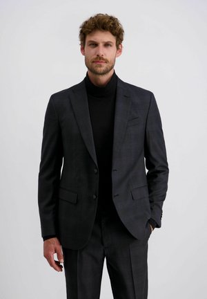 Mann mit lockigem Haar, der einen dunklen karierten Anzug und einen schwarzen Rollkragenpullover trägt, steht mit einer Hand in der Tasche vor einem weißen Hintergrund.