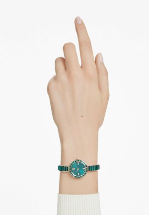 Montre ronde turquoise avec des accents dorés, présentant un bracelet assorti en maillons rectangulaires. Main levée, affichant le cadran de la montre et ses détails de design.