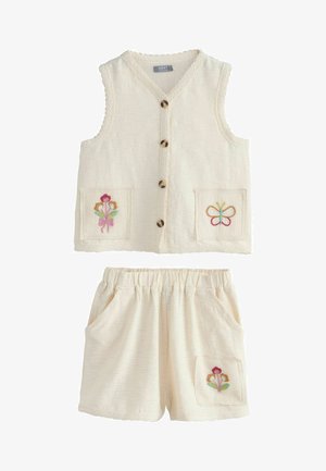 Bekvapis, kreminės spalvos liemenė be rankovių su gėlių ir drugelių siuvinėtais kišenėmis, derinama su tampriais šortais su elastine juosmens dalimi.