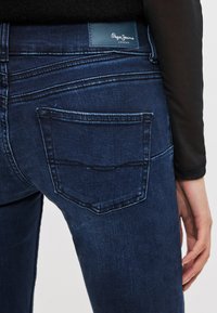Pantalones vaqueros de mezclilla azul oscuro con bolsillo trasero con costura curva, parche de cuero con la marca y textura suave. Vista en primer plano desde un lado.