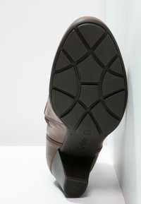 Bota de cuero marrón con una suela de goma negra texturizada que presenta un patrón de relieve. El tacón es moderadamente elevado y tiene detalles de costura sutiles.