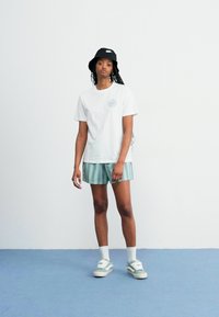 Hvit bomulls t-skjorte med et sirkulært logo, sammen med grønne stripete shorts. Brukt med en svart bøttehatt og grønne joggesko.
