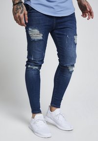 Man som bär trasiga, mörkblå skinny jeans och vita sportskor, stående mot en enkel ljus bakgrund.
