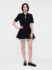 Femme aux cheveux courts et foncés, portant une robe noire à manches courtes avec une jupe évasée et un ourlet à volants blancs, associée à des chaussures Mary Jane noires.