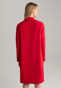 Rode jurk met lange mouwen, rechte silhouet en ronde kraag. Heeft een klein logodetail op de achterkant van de halslijn. Vloeiende textuur van de stof.