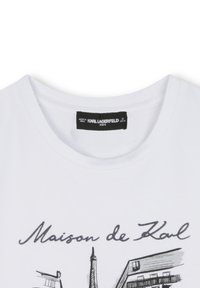T-shirt en coton blanc avec un col rond. Impression sérigraphique "Maison de Karl" et designs architecturaux en noir. Étiquette visible à l'intérieur.