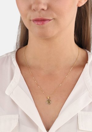 Collana d'oro con un pendente circolare raffigurante un design ad albero, indossata sopra una camicia bianca. La catena è sottile con piccoli anelli.