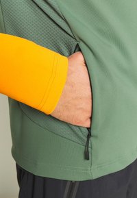 Groene jas van gestructureerde stof met een oranje mouw. Voorzien van een ritszak aan de zijkant en een strakke, gestroomlijnde vormgeving.