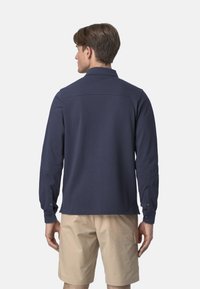 Camicia a maniche lunghe blu navy con colletto, polsini con bottone e orlo dritto. Texture liscia e design minimalista, abbinata a pantaloni corti beige.