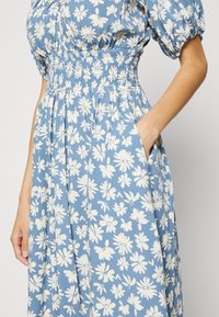 Blauwe bloemenjurk met witte madeliefjespatronen, gesmokte taille, korte pofmouwen en zijzakken voor extra functionaliteit.