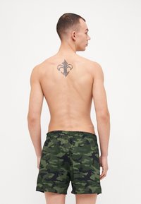 Shorts de bain camouflés en vert et noir avec une taille élastique. La vue arrière montre un tatouage et un design minimaliste sans détails supplémentaires.