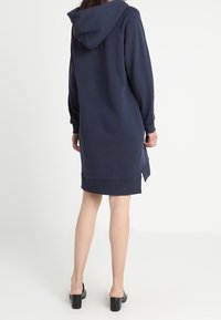 Granatowa sukienka z kapturem wykonana z miękkiego materiału, z kieszenią kangurką i ściągaczami na rękawach. Projekt jest długi, z rozcięciem po bokach na dole.