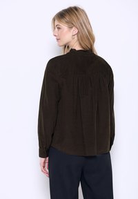 Camicia in corduroy marrone con maniche lunghe, colletto arricciato e dettagli di pieghe sul retro. Abbinata a pantaloni neri. Texture liscia.