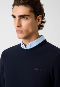 Marineblauwe gebreide trui met ronde hals, met daaronder een lichtblauw overhemd met kraag. Het woord "GUESS" is op de borst geborduurd.