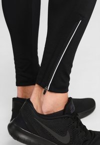 Leggings de sport noirs avec des ouvertures zippées aux chevilles, associés à des baskets noires. Le tissu semble lisse avec un fit ajusté.