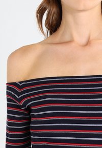 Top de hombros descubiertos a rayas en azul marino, rojo, gris y blanco. Tejido suave y elástico con rayas horizontales y un diseño ajustado.