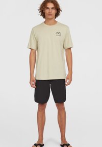 Lichtbeige katoenen t-shirt met korte mouwen en een geprinte opdruk op de borst, gecombineerd met zwarte shorts en zwarte sandalen.