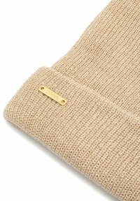 Beżowa dzianinowa czapka z wełny z zawiniętym mankietem i małą złotą metalową metką marki przymocowaną do mankietu.