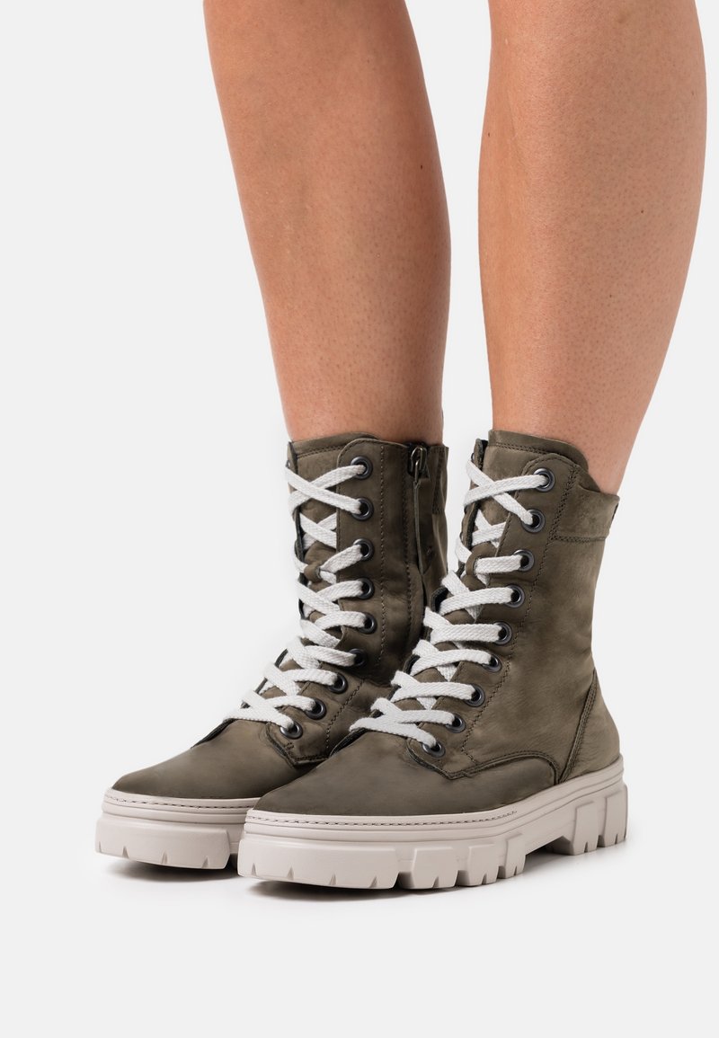 Bottes de combat vert olive usées à lacets avec semelles blanches épaisses et fermeture éclair latérale, montrées sur des jambes nues sur fond blanc.