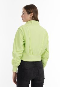 Giacca bomber verde neon con colletto alto, vita elastica e maniche voluminose, indossata con jeans neri. Finitura liscia e lucida.