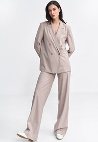 Beżowy garnitur w paski z podwójnym zapięciem na marynarce, brązowymi guzikami i szerokimi spodniami, w połączeniu z białymi butami. Gładka tekstura.