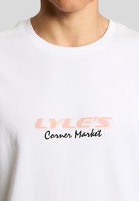 Κοντινό πλάνο ενός ατόμου που φοράει λευκό μπλουζάκι με τη λέξη "LYLE'S" σε ανοιχτό ροζ και το κείμενο "Corner Market" σε μαύρη καλλιγραφική γραφή στο μπροστινό μέρος.