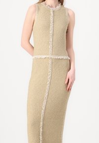 Robe en tricot beige avec une finition texturée, design sans manches, bords effilochés le long des coutures, affichant une silhouette profilée.