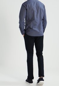 Mężczyzna w niebieskiej koszuli z długim rękawem, czarnych spodniach i czarnych trampkach z białymi podeszwami, stojący z rękami w kieszeniach, zwrócony plecami.