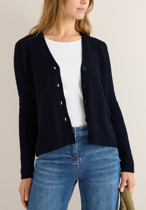 Navy Cardigan aus weichem Strickstoff, mit V-Ausschnitt, Knopfverzierungen vorne und gerippten Bündchen und Saum. Getragen über einem weißen Hemd.